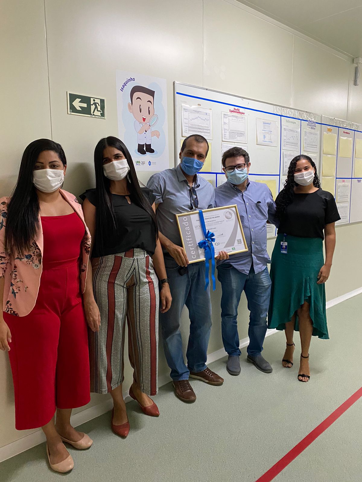 Cinco pessoas, todas de máscara, estão de pé em um corredor hospitalar, um ao lado do outro. A pessoa que está no meio, segura uma placa com um laço azul.